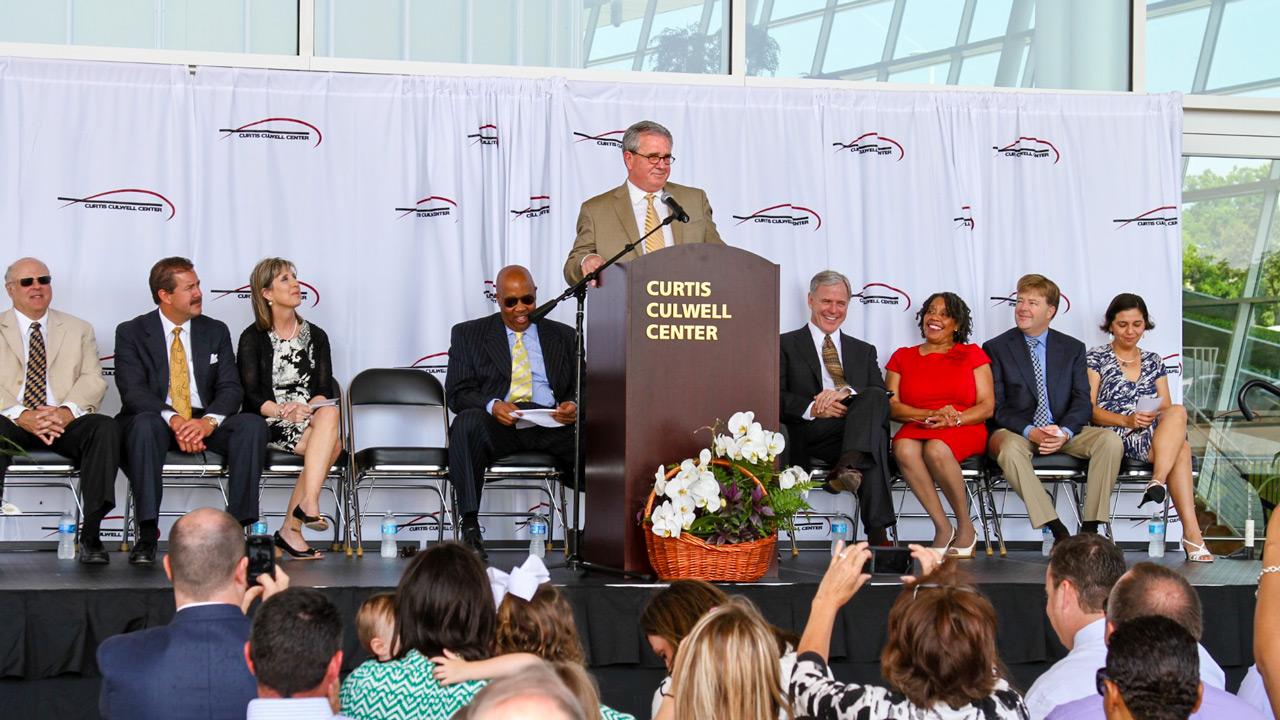 Dr. Culwell gives a speech on stage at the CCC Naming Ceremony. The Garland ISD Board of Trustees and a crowd look on, smiling and taking photos.