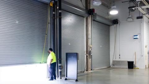 Three large loading bay doors lead into a concreted area fitted with docking technology. A security guard looks out one of the loading doors that is ajar.