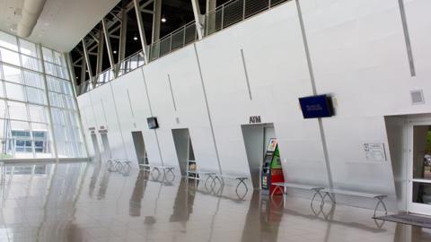 Light from glass walls illuminates the tiled floor of the lower concourse, featuring a modern slanted wall, lined with benches, an ATM, and screens.