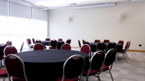 A conference room featuring a wall of windows, a projector screen, LED lighting, and round tables with chairs.