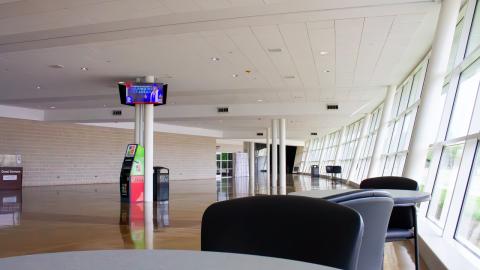 A large wall of glass illuminates a large space with polished floors in natural light. Tables and seats sit among the edge, with an ATM, screen TV, and vending machine visible.