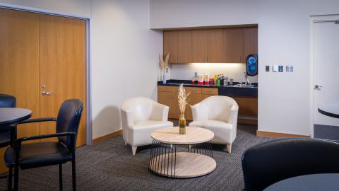 Cream colored chairs surround a modern wood and metal coffee table. Behind them is a small kitchenette, and high tables with chairs sit in front.