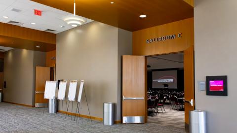 A wide view of the wood-paneled entrances to the ballroom, featuring pendant chandeliers, recessed lighting, and digital event title screens.