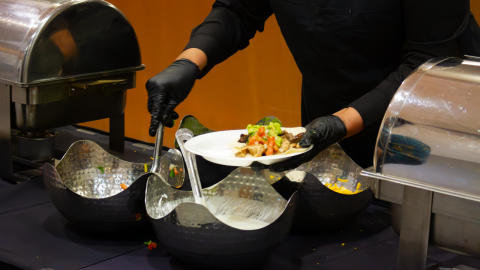 A caterer serves food on to a plate from patterned metal bowls. The sleek serving containers sit on a black table cover.