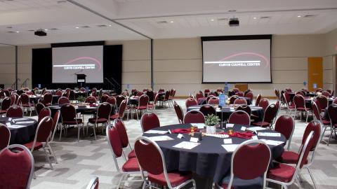 A wide view of the CCC ballroom, featuring multiple projectors with screens, a stage and podium, sound equipment and nice round tables with chairs.