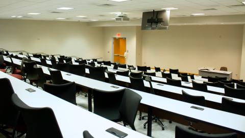 A auditorium-style lecture hall filled with seminar tables and chairs, featuring a podium, projector, and presenter notes screen.