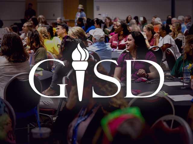 From afar, we see a woman smiling at a presenter. She sits in a crowd, everyone seated at round tables for a lecture. The letters "GISD" are superimposed on the image.