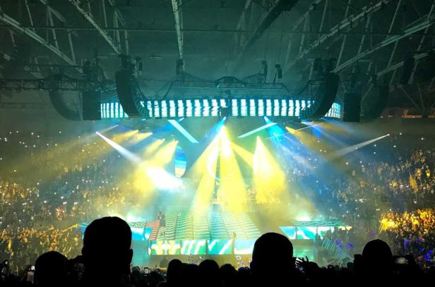 Overlooking the arena stage from upper seating. The stage has bright yellow, blue and aqua lighting.