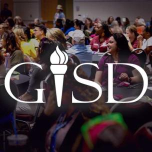 From afar, we see a woman smiling at a presenter. She sits in a crowd, everyone seated at round tables for a lecture. The letters "GISD" are superimposed on the image.